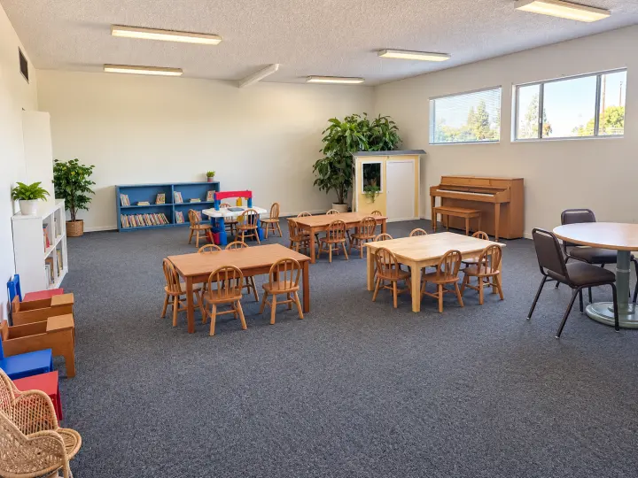 Preschool classroom with small wooden tables and chairs, bookshelves, plants and an upright piano