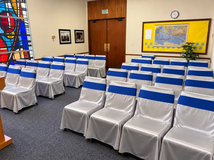 Side view of rows of covered chairs with blue sashes beside a stained-glass window